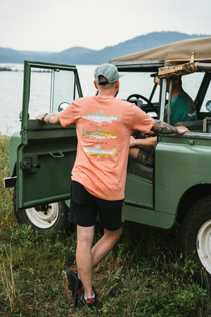 Coral short-sleeve t-shirt with colorful block print trout design worn with black shorts and gray cap by green vintage.